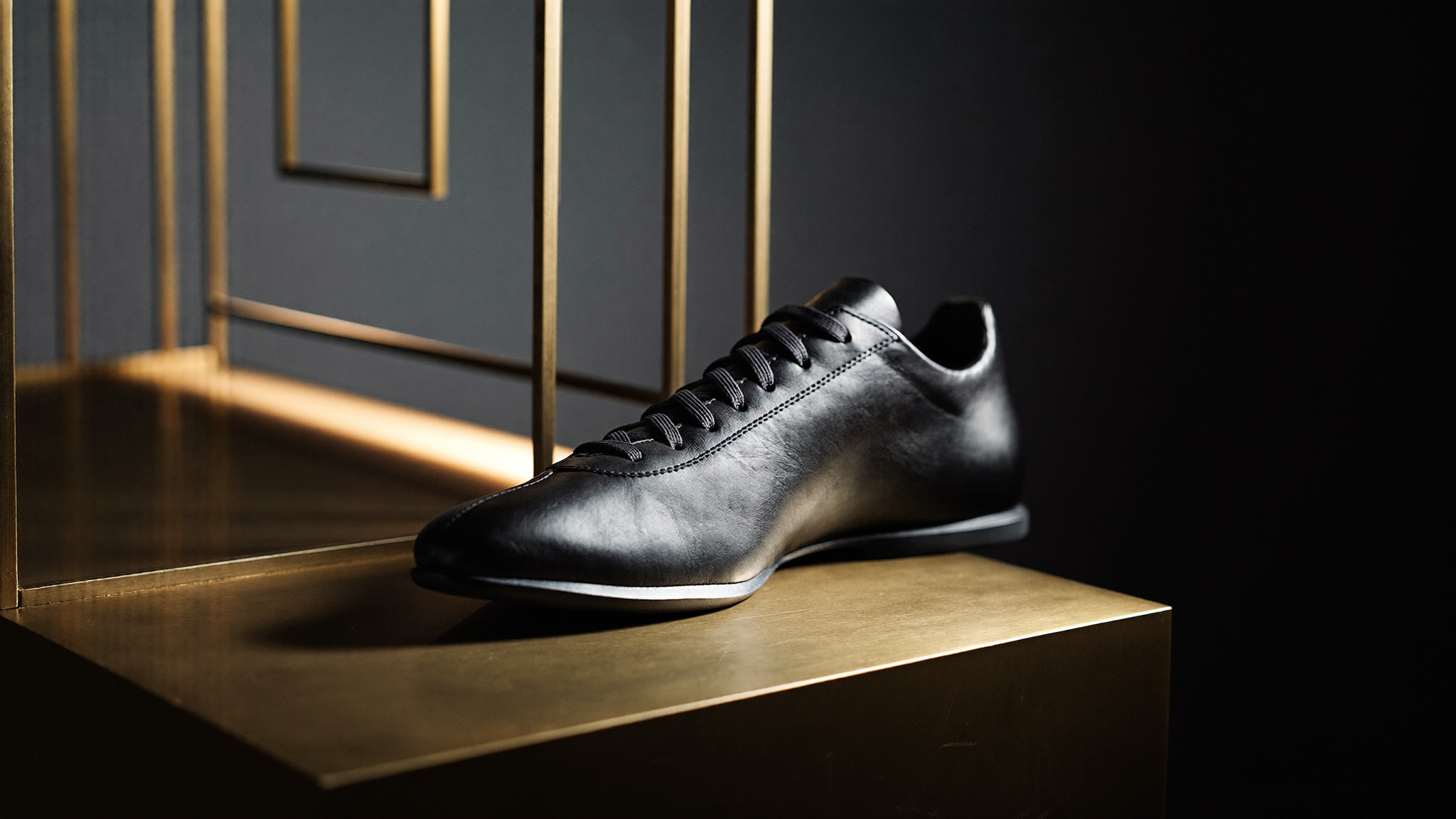 Ein einzelner Ledersneaker, diagonal auf einer schrägen Messingplinthe platziert, im kinematischen Streiflicht vor dunklem Studiohintergrund