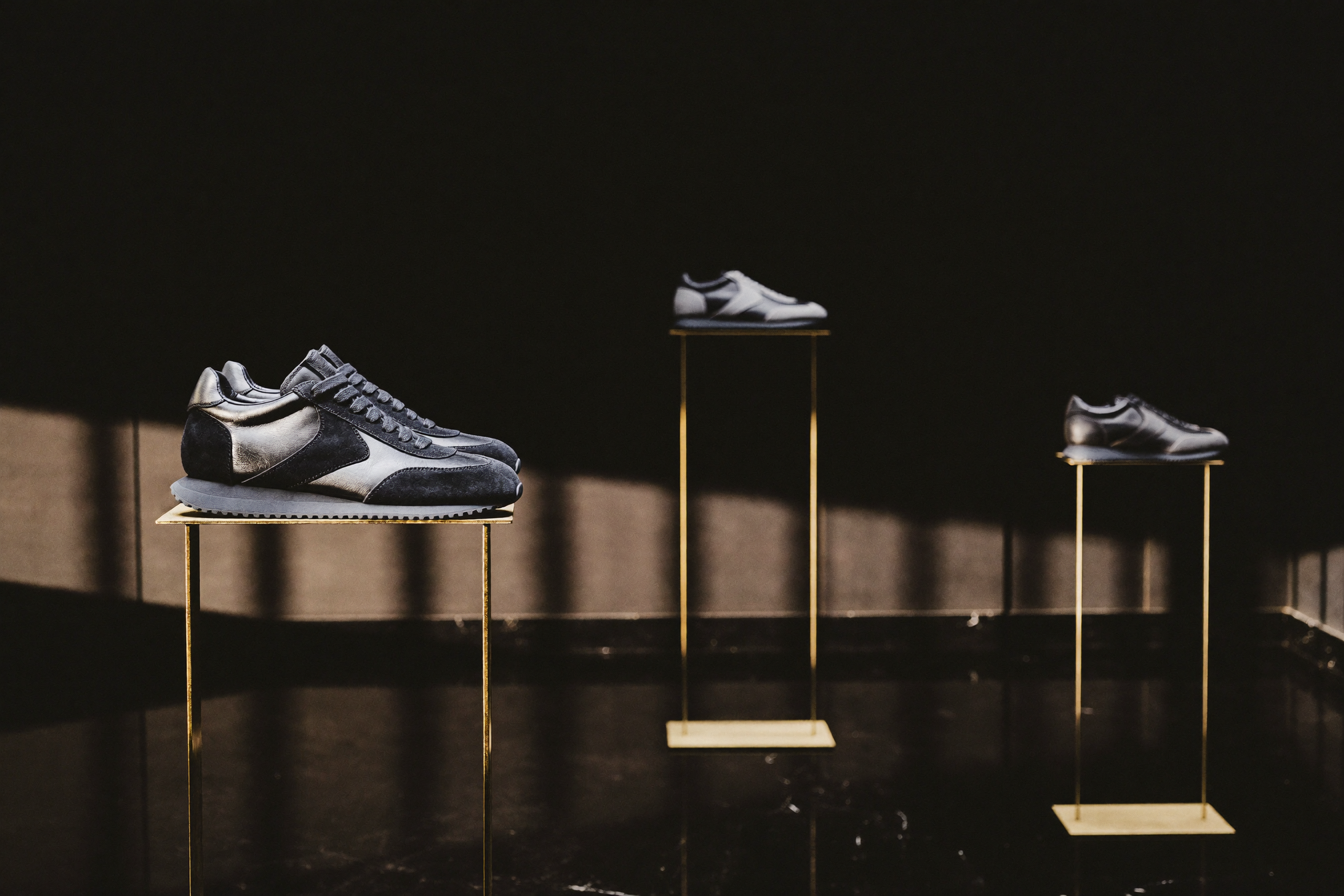 Drei Paar Sneakers, asymmetrisch auf schmalen Messingschienen präsentiert, mit dramatischem Streiflicht im Atelier